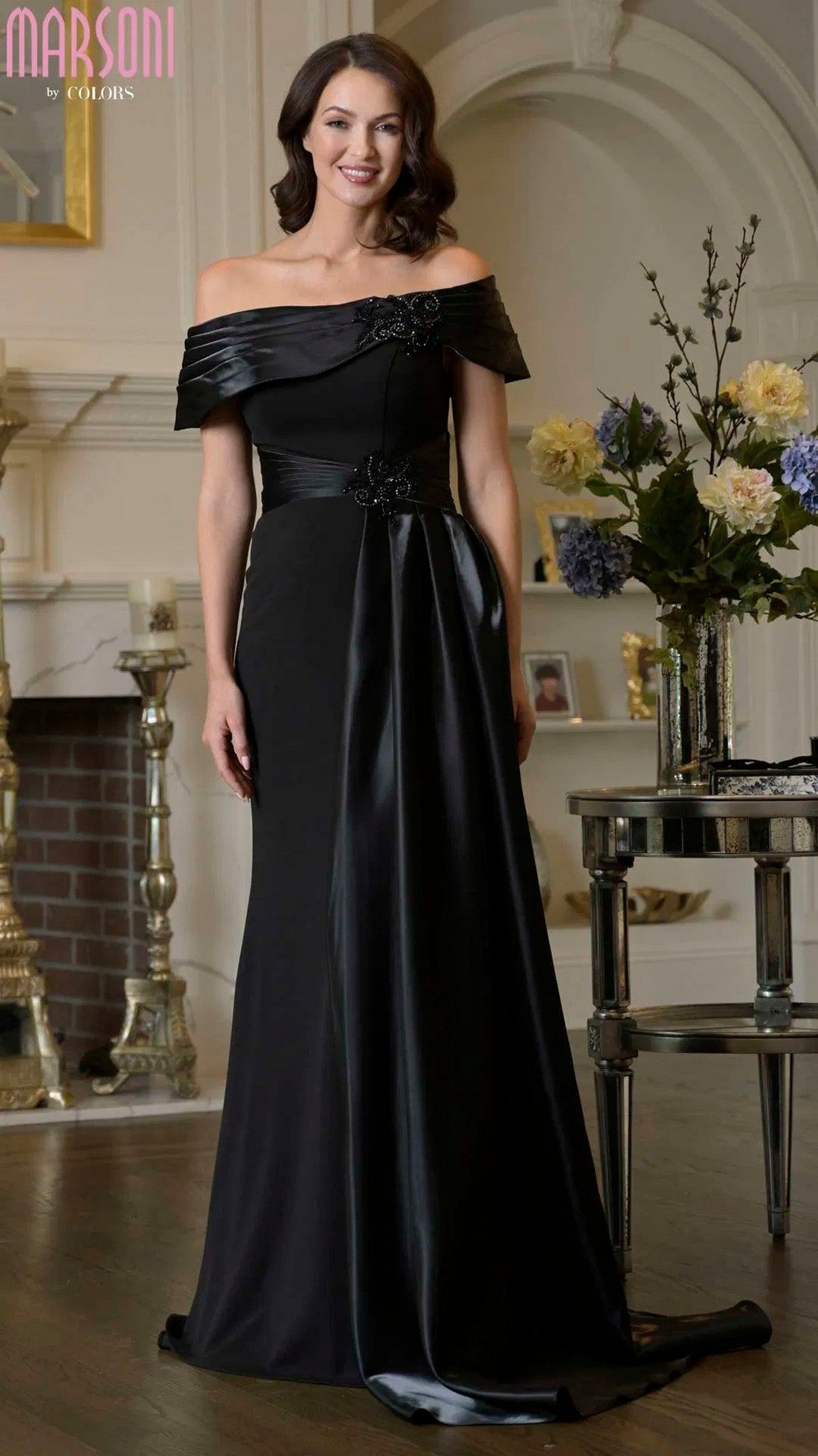 A woman stands indoors in the Marsoni M347, a floor-length black formal dress with floral embellishments and a sheath silhouette, next to a table with a floral arrangement.